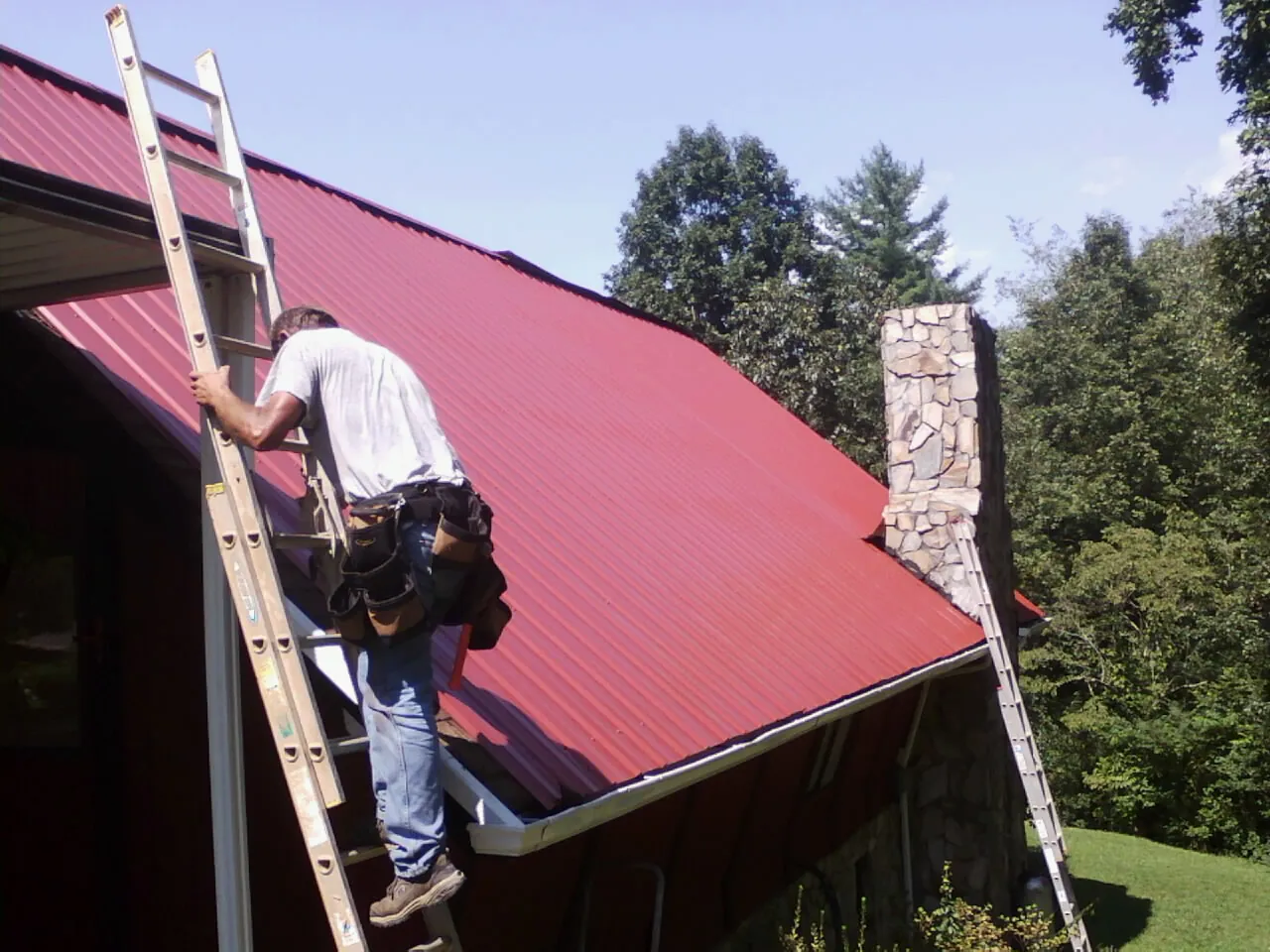 Roofer on a ladder beside a red metal roof and stone chimney — Soffit & Fascia Repair by Roofing Brothers in Lehighton